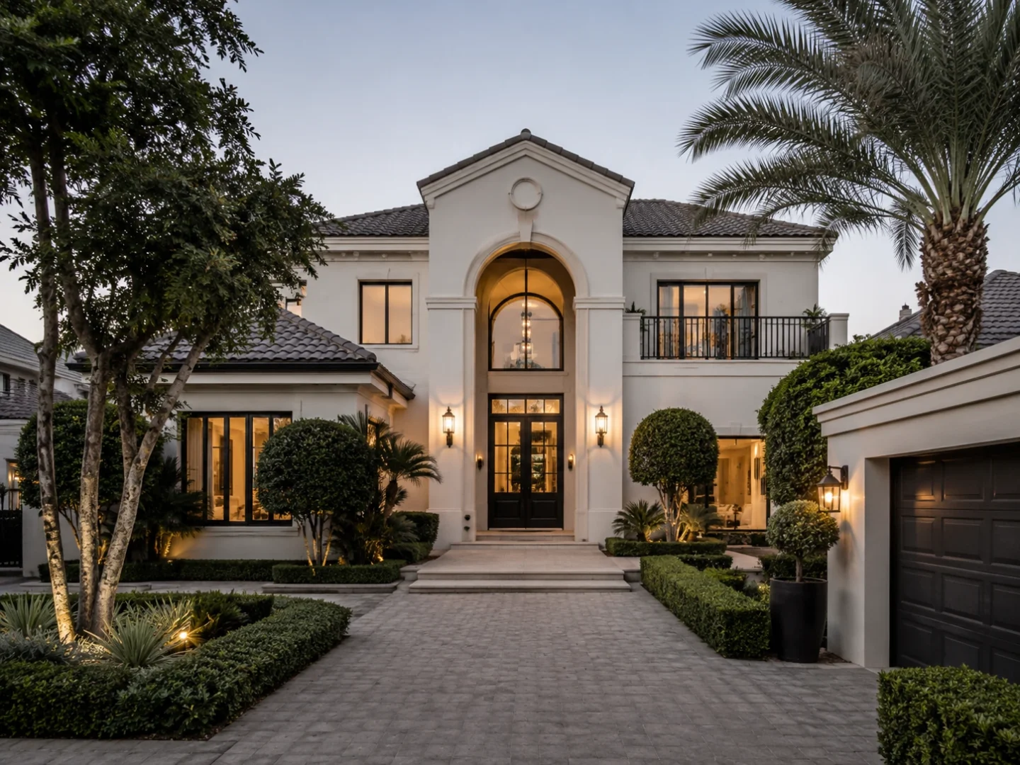 A grand villa with an arched entrance glowing warmly at early evening, framed by manicured hedges and mature palms