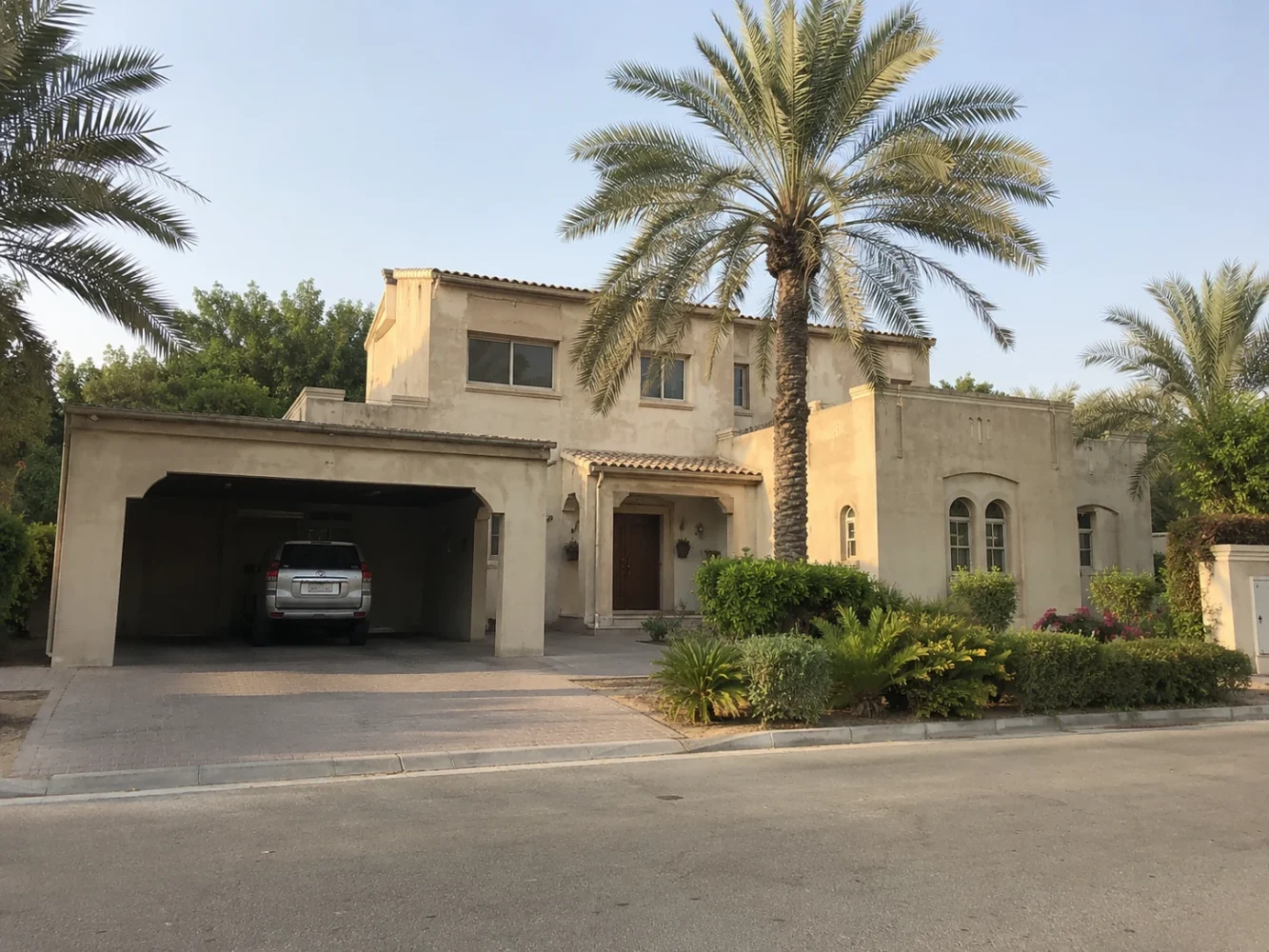 An older two-storey Dubai villa with a covered carport and mature palms on a quiet residential street, showing clear upgrade potential