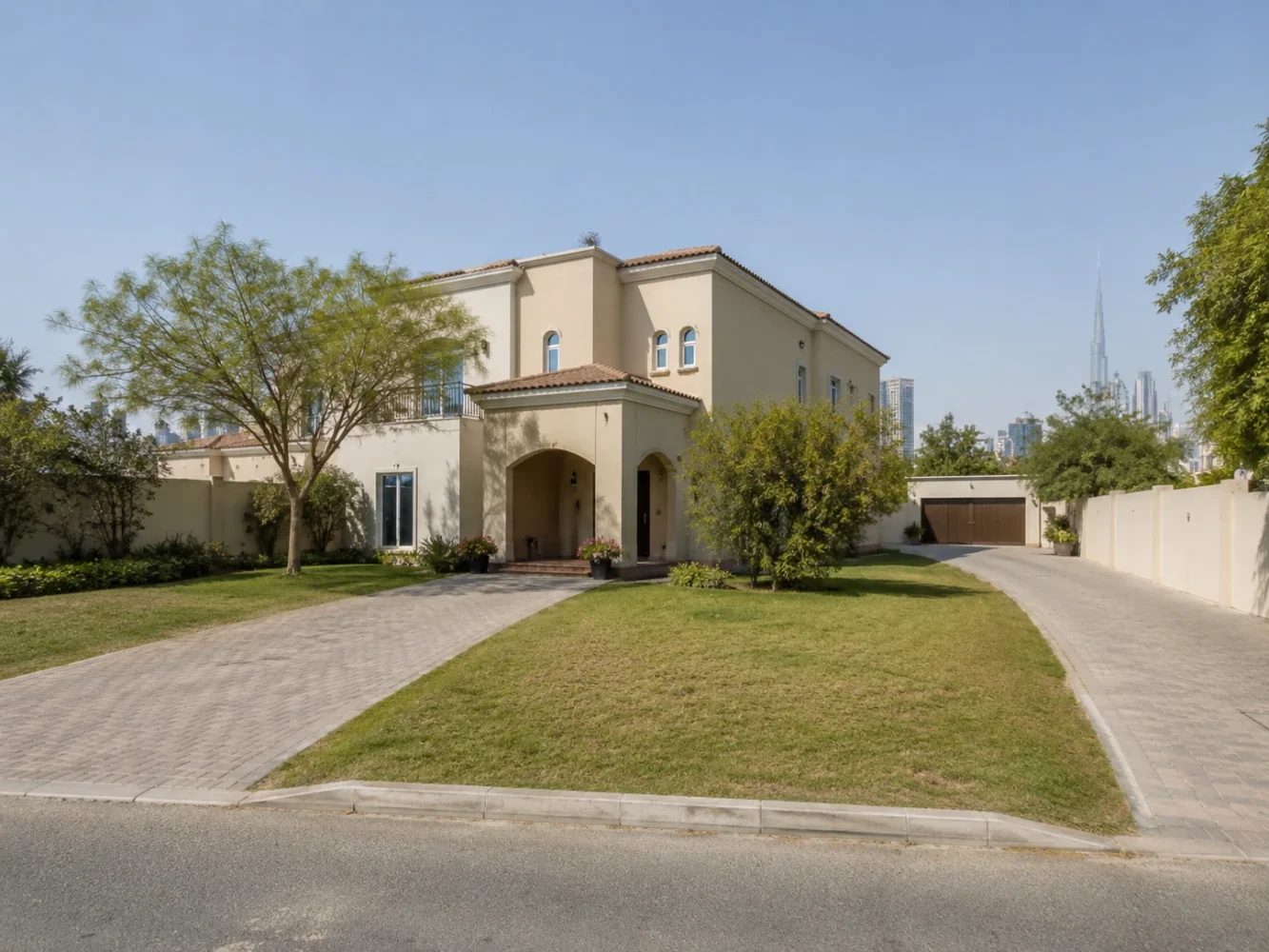 A two-storey Dubai villa on a large grassed corner plot with driveways and mature trees, with the city skyline behind